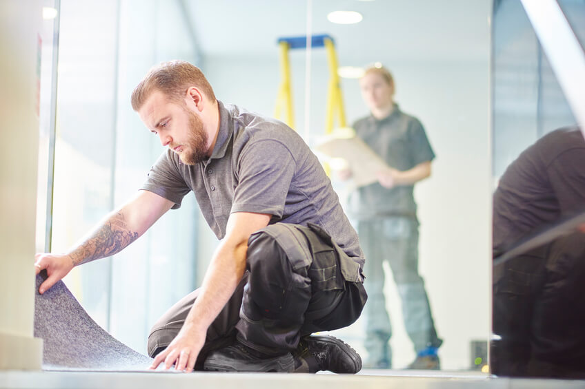 male carpet fitter fitting office carpet tiles a male carpet fitter is installing carpet in a modern office refurbishment. IN the background a female co-worker can be seen holding plans . They are wearing safety workwear.